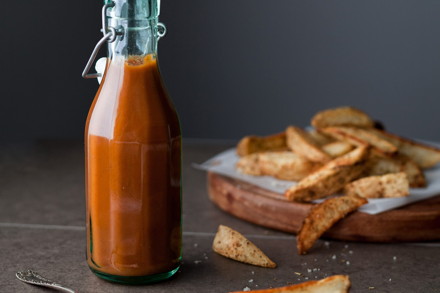 Una botella de cristal con ketchup casero con patatas al fondo.